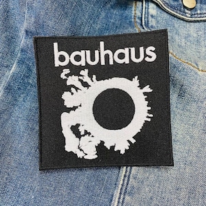 May include: Black square patch with the word "bauhaus" in white, above a white graphic with a black circle in the center. The patch is sewn onto a denim jacket.