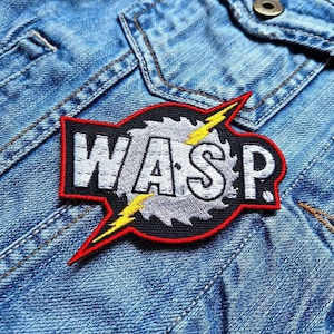 May include: Embroidered patch with the word "WASP" in white, outlined in black and red. The design includes a saw blade and yellow lightning bolts. The patch is sewn onto a blue denim jacket.