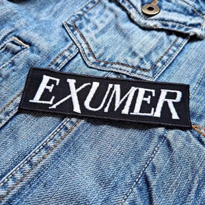 May include: A black rectangular patch with the word "EXUMER" embroidered in white, attached to a light blue denim jacket. The jacket shows stitching and a button.