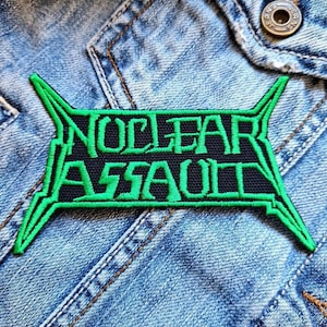 Puede incluir: Un parche negro con las palabras "NUCLEAR ASSAULT" bordadas en verde. El parche tiene un borde verde con puntas angulares. El parche está cosido en una chaqueta vaquera.