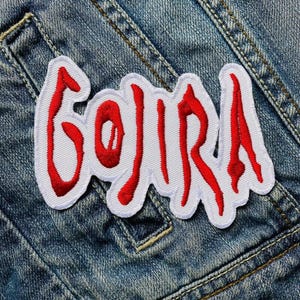 Könnte beinhalten: Weißer gestickter Aufnäher mit dem Wort "GOJIRA" in roten Buchstaben. Der Aufnäher ist auf eine blaue Jeansjacke genäht.