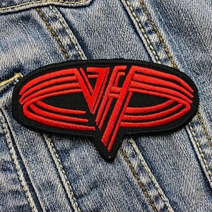Pode incluir: Patch bordado com o logotipo vermelho e preto de Van Halen. O patch tem uma forma oval preta com linhas vermelhas e as letras "VH" em uma fonte estilizada. O patch é costurado em uma jaqueta jeans.