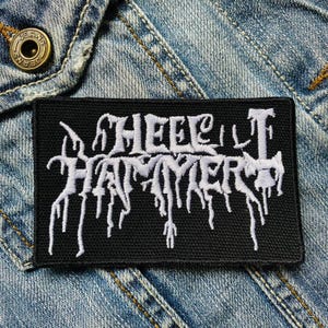 Op de afbeelding: Een zwart rechthoekig embleem met de wit geborduurde tekst "HEEL HAMMER" in een gotisch lettertype. De patch is geplaatst tegen een blauwe denim achtergrond. Een knoop is zichtbaar in de linkerbovenhoek.