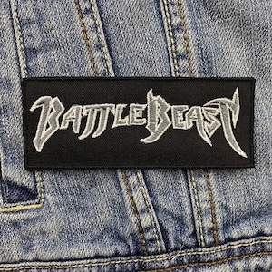 May include: Black patch with white embroidered text that reads "BATTLE BEAST" on a denim jacket.