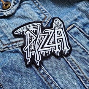 Könnte beinhalten: Ein schwarz-weißer, gestickter Aufnäher mit dem Wort "PIZZA" in einer stilisierten Schriftart. Der Aufnäher ist auf einer Jeansjacke befestigt. Das Design umfasst eine Sense, Spinnweben und tropfende Details.