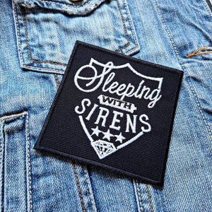 May include: Black and white embroidered patch with the text "Sleeping with Sirens" in a shield shape with three stars and a diamond.