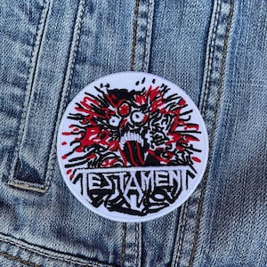 May include: White and red embroidered patch with a black and white illustration of a screaming face with the word "TESTAMENT" written below it.