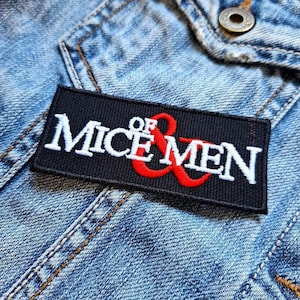 May include: Black and white embroidered patch with the text "Of Mice & Men" in white and a red ampersand symbol. The patch is sewn onto a blue denim jacket.
