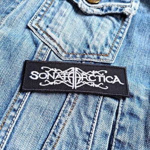 May include: A black rectangular patch with white embroidered text that reads "SONATA ARCTICA" in a stylized font. The patch is sewn onto a light blue denim jacket, with visible seams and a pocket.