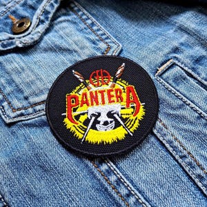 May include: A round, black patch featuring the word "PANTERA" in red and yellow lettering. The patch includes a skull design and crossed weapons, with a yellow and white border. The patch is sewn onto a denim jacket.