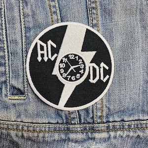 以下が含まれることがあります： 白と黒の刺繍パッチ。稲妻の白い稲妻の両側に「AC」と「DC」の文字が配置されています。中央には時計の文字盤があります。パッチはデニムジャケットに縫い付けられています。