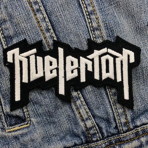 Può includere: Toppa ricamata in bianco e nero con la parola "Tuelertown" in un font stilizzato.