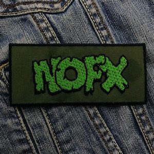 May include: Green embroidered patch with the word "NOFX" on a dark green background. The patch is on a blue denim jacket.