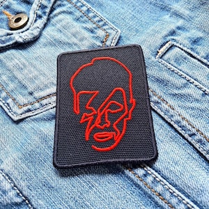 Könnte beinhalten: Ein schwarzer Patch mit einer roten Umrandung eines menschlichen Gesichts. Das Gesicht ist stilisiert und hat ein geometrisches Design.