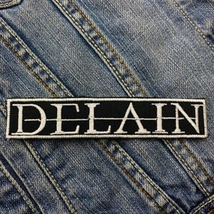 May include: Black and white embroidered patch with the word "DELAIN" in white lettering on a black background.
