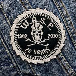 May include: A black and white embroidered patch with a skull and crossbones design. The text "W.A.S.P." is above the skull and the text "1982 28 Years 2010" is below the skull.