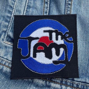 Puede incluir: Parche bordado negro con un diseño de diana blanco y azul y el texto "The Jam" en blanco.