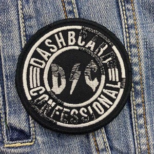 May include: A round, black embroidered patch with white text and a lightning bolt design. The text reads "DASHBLAST PROFESSIONAL" in a circular pattern around the central graphic. The patch is sewn onto a denim jacket.