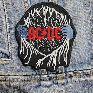 Op de afbeelding: Zwarte geborduurde patch met de bandnaam "AC/DC" in het rood. Het ontwerp bevat witte bliksemschichten en blauwe accenten, genaaid op een spijkerjasje. Een accessoire met rockmuziekthema.