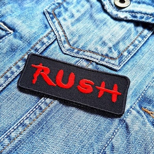 Könnte beinhalten: Ein schwarzer Stoff-Patch mit dem Wort "RUSH" in roten Buchstaben. Der Patch ist auf eine blaue Jeansjacke genäht.