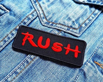 Peut inclure: Un écusson en tissu noir avec le mot "RUSH" en lettres rouges. L'écusson est cousu sur une veste en jean bleue.