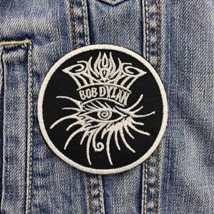May include: Black and white embroidered patch with a stylized eye and the text "Bob Dylan" in a banner.