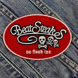 Könnte beinhalten: Ovaler, roter, gestickter Aufnäher mit den Worten "Beat Steakes" in weißer Schrift, mit Totenkopf- und gekreuzten Knochen-Grafiken. Der Text "no flesh inc" steht darunter. Der Aufnäher befindet sich auf einem Jeans-Hintergrund.