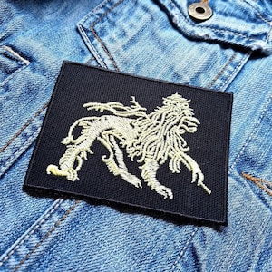 May include: A black rectangular patch with a cream-colored lion design, sewn onto a blue denim jacket. The lion is depicted in a detailed, stylized illustration. The patch is centered on the jacket, showcasing the embroidered artwork.
