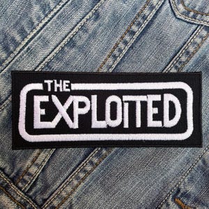 Pode incluir: Etiqueta bordada em preto e branco com o texto "THE EXPLOITED" em um retângulo branco com contorno preto. A etiqueta está em um fundo de jeans azul.