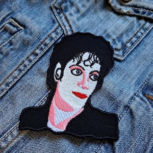 May include: Embroidered patch featuring a portrait of a person with black hair, red lips, and pink accents on a black background. The patch is sewn onto a denim jacket.