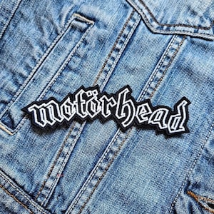 May include: A black and white embroidered patch with the word "Motorhead" in a stylized font. The patch is sewn onto a blue denim jacket.