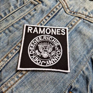 Puede incluir: Parche bordado en blanco y negro con la palabra "RAMONES" y un círculo que contiene los nombres "JOHNNY, DEE DEE, TOMMY y RICHIE" en un diseño circular.