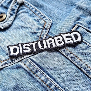 Puede incluir: Un parche bordado en blanco y negro con la palabra "DISTURBED" en letras blancas sobre un fondo negro. El parche está cosido en una chaqueta vaquera azul.
