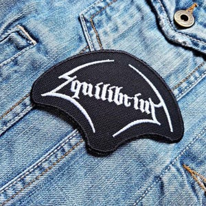 Könnte beinhalten: Schwarzer Aufnäher mit weiß gesticktem Text „Equilibrium“ in einer stilisierten Schriftart. Der Aufnäher ist auf eine hellblaue Jeansjacke genäht. Der Aufnäher hat eine gebogene Form.