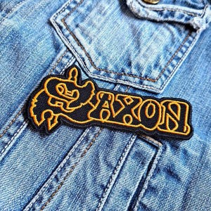 Könnte beinhalten: Ein gestickter schwarzer und gelber Aufnäher mit dem Wort "SAXON" in einer stilisierten Schrift.