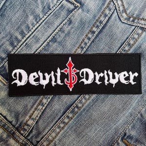Puede incluir: Parche bordado en blanco y negro con el texto "Devil Driver" en fuente gótica. Una cruz roja está centrada entre las palabras.
