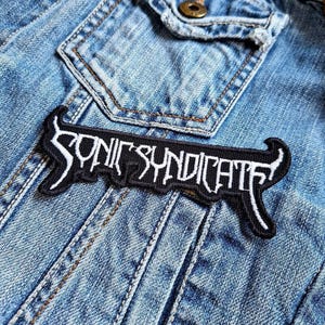 May include: Black and white embroidered patch with the text "Sonic Syndicate" on a denim jacket.