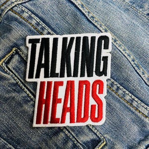 Op de afbeelding: Een geborduurde patch in zwart en wit met de woorden "Talking Heads" in rode en witte letters. De patch is op een blauwe denim achtergrond.
