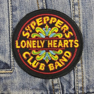 Puede incluir: Parche redondo bordado con las palabras "Sgt. Pepper's Lonely Hearts Club Band" en letras amarillas. El parche tiene un fondo negro con detalles rojos y azules, sobre una chaqueta vaquera.