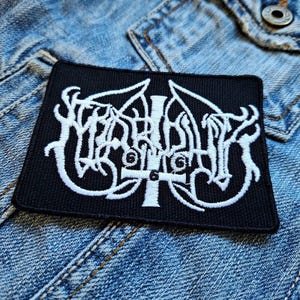 Op de afbeelding: Een zwarte geborduurde patch met een wit logo van de band "Watain". Het logo toont een gestileerd kruis met een schedel en vleugels.
