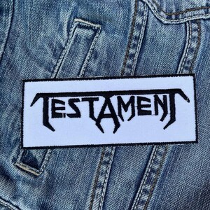 May include: White patch with black embroidered text "TESTAMENT" on a blue denim jacket.