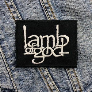 Könnte beinhalten: Schwarzer rechteckiger Aufnäher mit dem weißen Text "Lamb of God". Der Aufnäher befindet sich auf einer Jeansjacke. Der Text ist in einer stilisierten Schriftart.