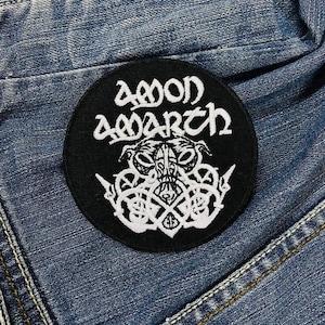 Könnte beinhalten: Ein schwarzer, runder Aufnäher mit dem weißen Schriftzug "AMON AMARTH". Darunter befindet sich ein weißes, detailliertes Design mit einer gehörnten Figur und Knotenmustern. Der Aufnäher liegt auf einem Jeansstoff-Hintergrund.