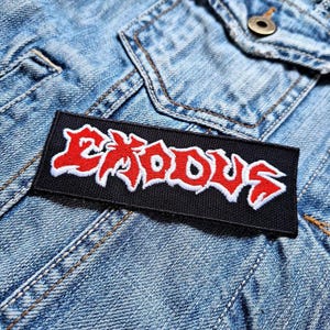 Può includere: Una toppa nera con cuciture bianche che presenta la parola "EXODUS" in lettere rosse. La toppa è su una giacca di jeans blu.