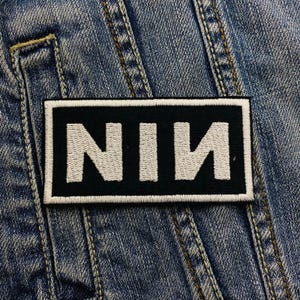 Pode incluir: Um patch retangular preto com as letras "NIN" bordadas em branco. O patch tem uma borda branca e é costurado em uma jaqueta jeans. A jaqueta jeans tem costuras visíveis.