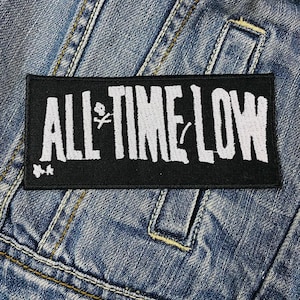 Puede incluir: Un parche rectangular negro con la inscripción blanca "ALL TIME LOW". El parche incluye un símbolo de calavera y huesos cruzados y un diseño floral. Está cosido a una chaqueta vaquera azul.