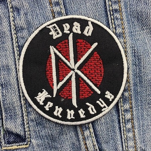 Op de afbeelding: Een ronde zwarte patch met de woorden "Dead Kennedys" in witte letters. De patch heeft een rood-wit grafisch ontwerp in het midden en is op een spijkerjas genaaid.