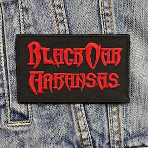 Pode incluir: Um patch retangular preto com as palavras "Black Oak Arkansas" bordadas em vermelho. O patch é apresentado em um fundo de ganga, destacando o contraste entre as cores e as texturas.