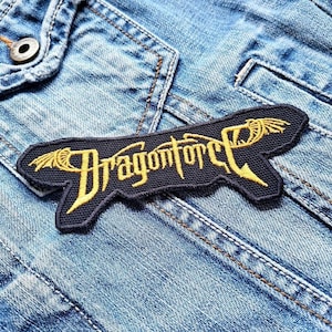Pode incluir: Um remendo bordado preto e dourado com o texto "Dragonforce" em uma jaqueta de jeans.