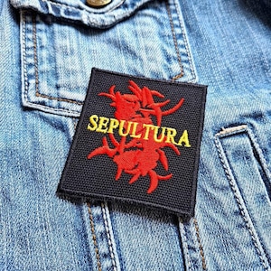May include: A black fabric patch with a red and yellow embroidered design. The design features a stylized logo with the word "SEPULTURA" in yellow lettering.